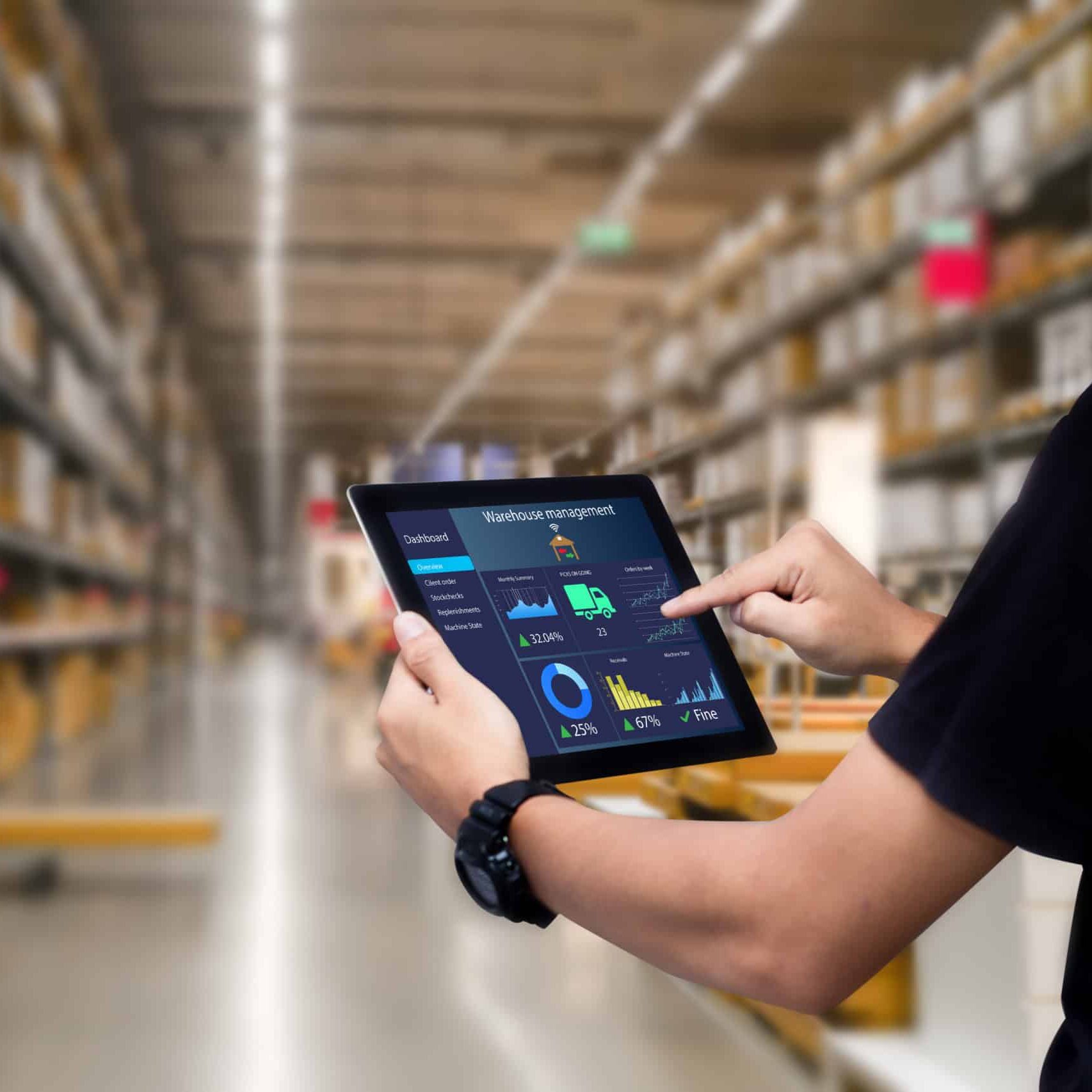 A supervisor stands in a warehouse, holding a smart device and reviewing a warehouse management app for inventory tracking and logistics monitoring.